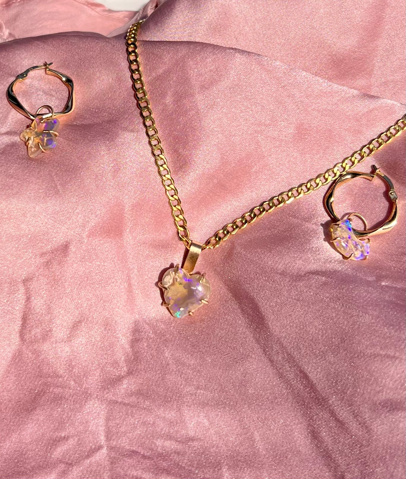 Gold necklace with a heart-shaped opal pendant and matching solid gold slight twist earrings on a pink fabric background